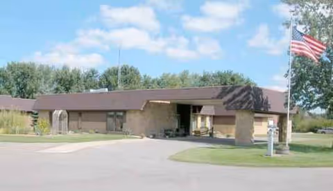 Single-story brick senior living facility with entrance, flagpole, and parking lot.