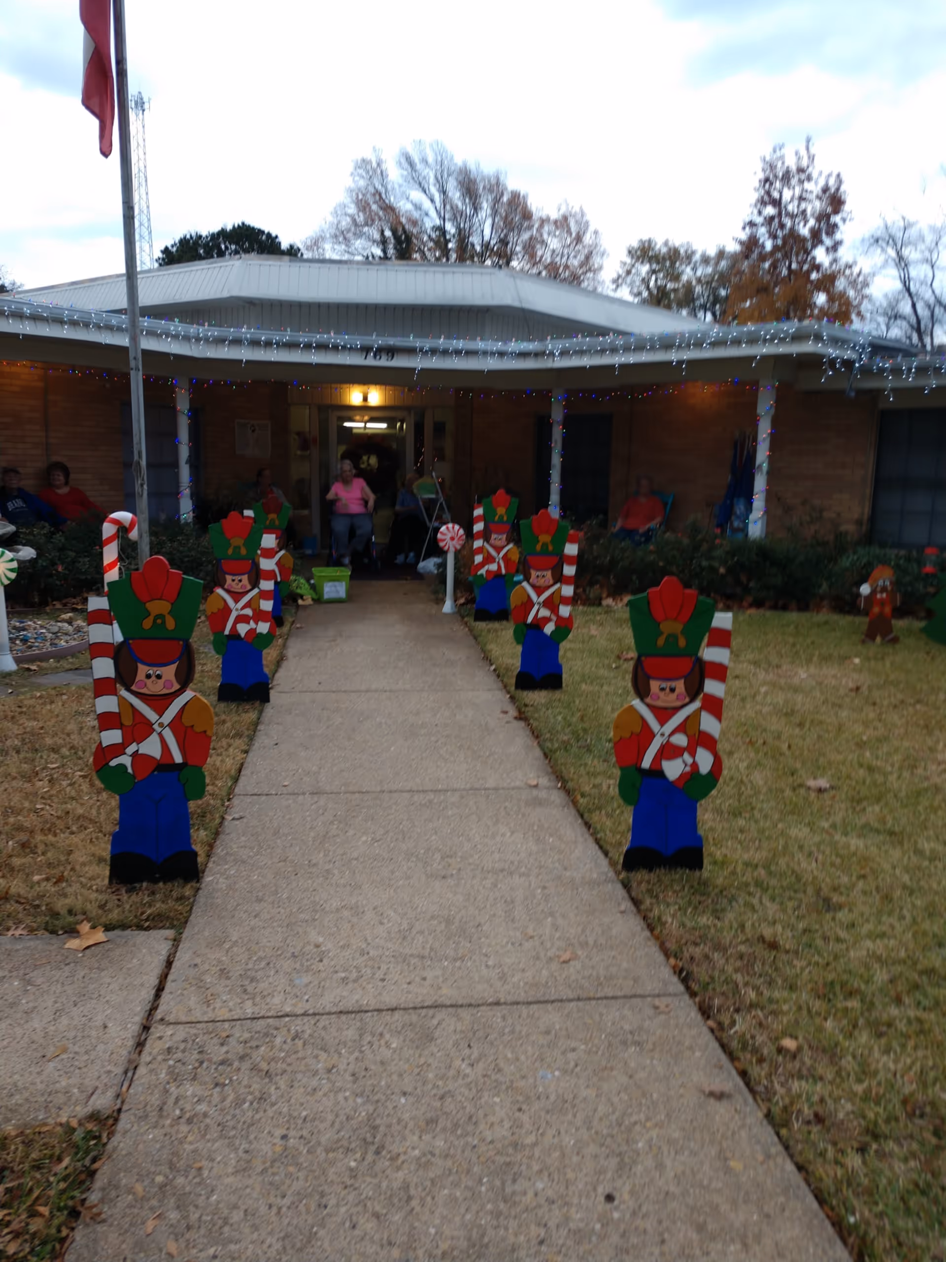 Outdoor walkway leading to the entrance of a single-story brick building decorated with Christmas lights and wooden toy soldier decorations lining both sides of the path. Several people are sitting near the entrance under a covered porch.