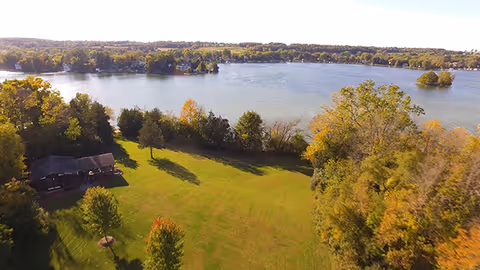 Aerial view of a lakeside property showing a grassy lawn, scattered trees, and a small building beside a large lake.