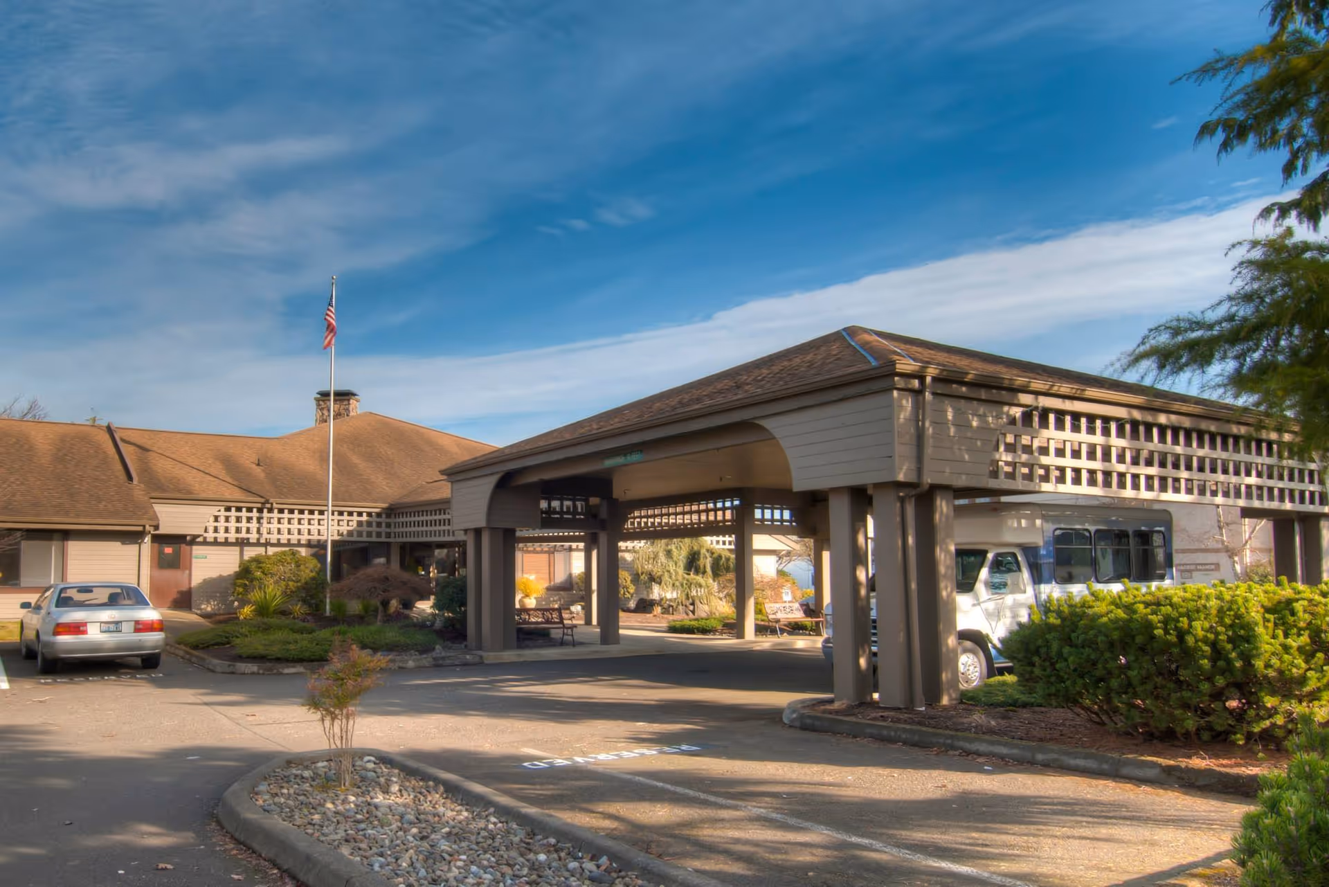 Exterior view of Channel Point Village senior living facility showing a covered entrance with a driveway, a parked car, a shuttle bus, landscaped bushes, and an American flag on a flagpole under a partly cloudy blue sky.