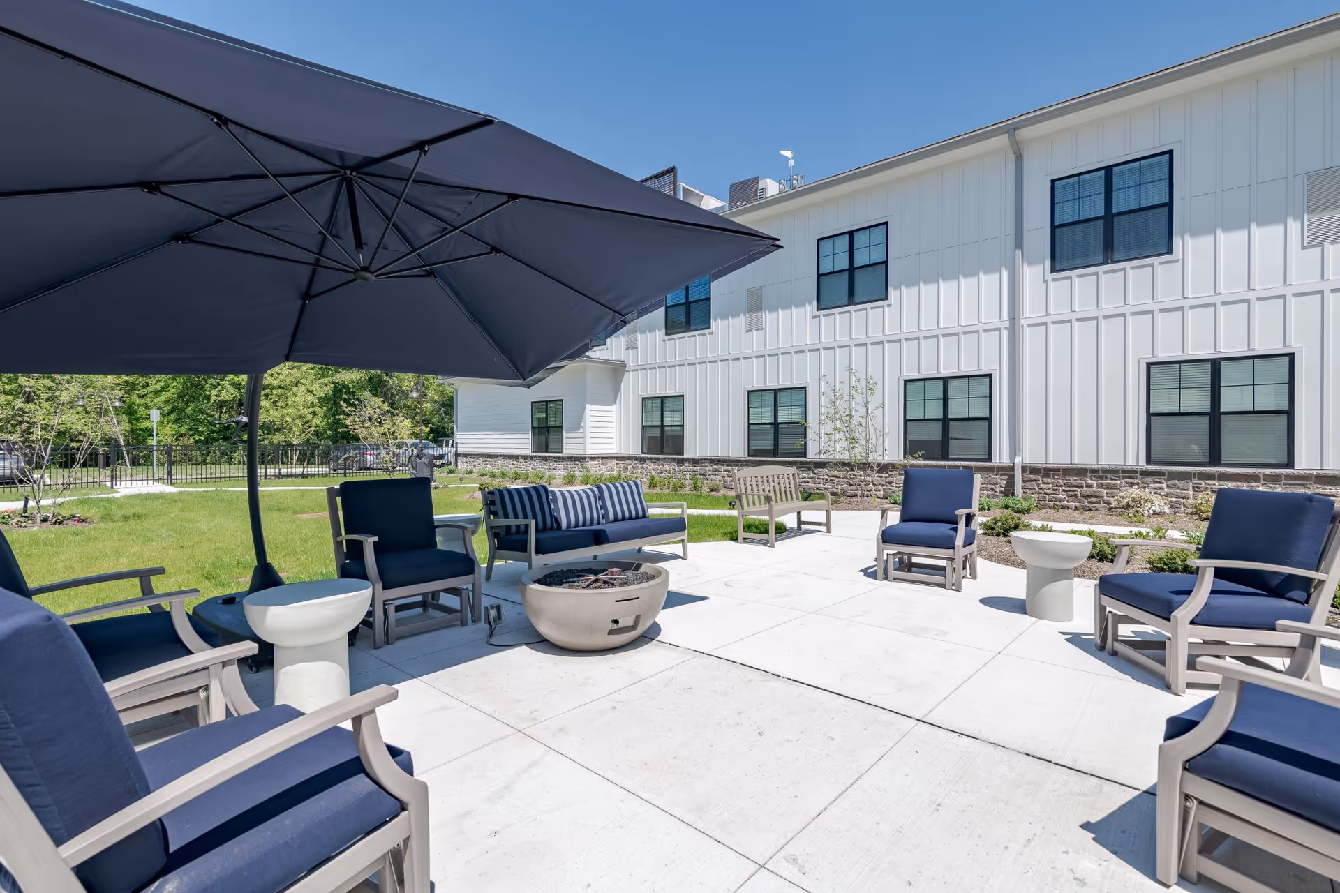 Outdoor patio area with cushioned chairs arranged around a fire pit, a large umbrella providing shade, and a white building with multiple windows in the background.