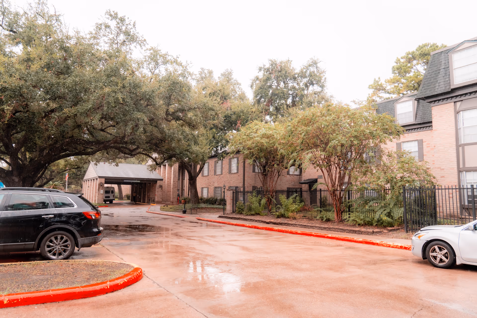 Driveway and front entrance of a brick senior living building with parked cars and large oak trees.