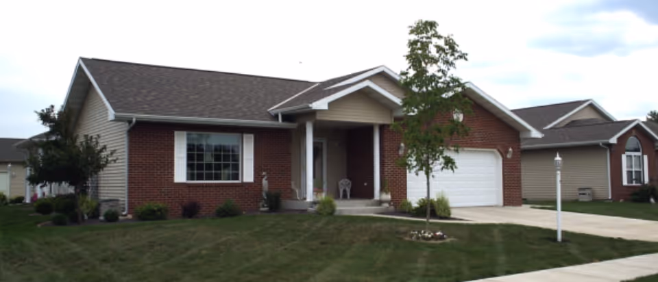 Single-story residential building with a brick and siding exterior, a front porch with two chairs, a garage, a small tree, and a well-maintained lawn.