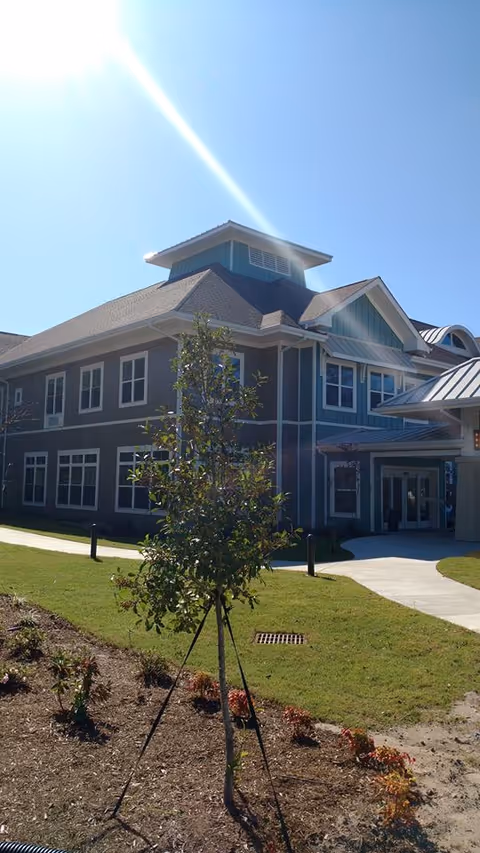 Exterior view of a two-story building with blue and gray siding, multiple windows, and a peaked roof under a clear blue sky with sunlight shining. In the foreground, there is a small tree supported by stakes and a landscaped area with grass and small plants. A curved concrete walkway leads to the building entrance.