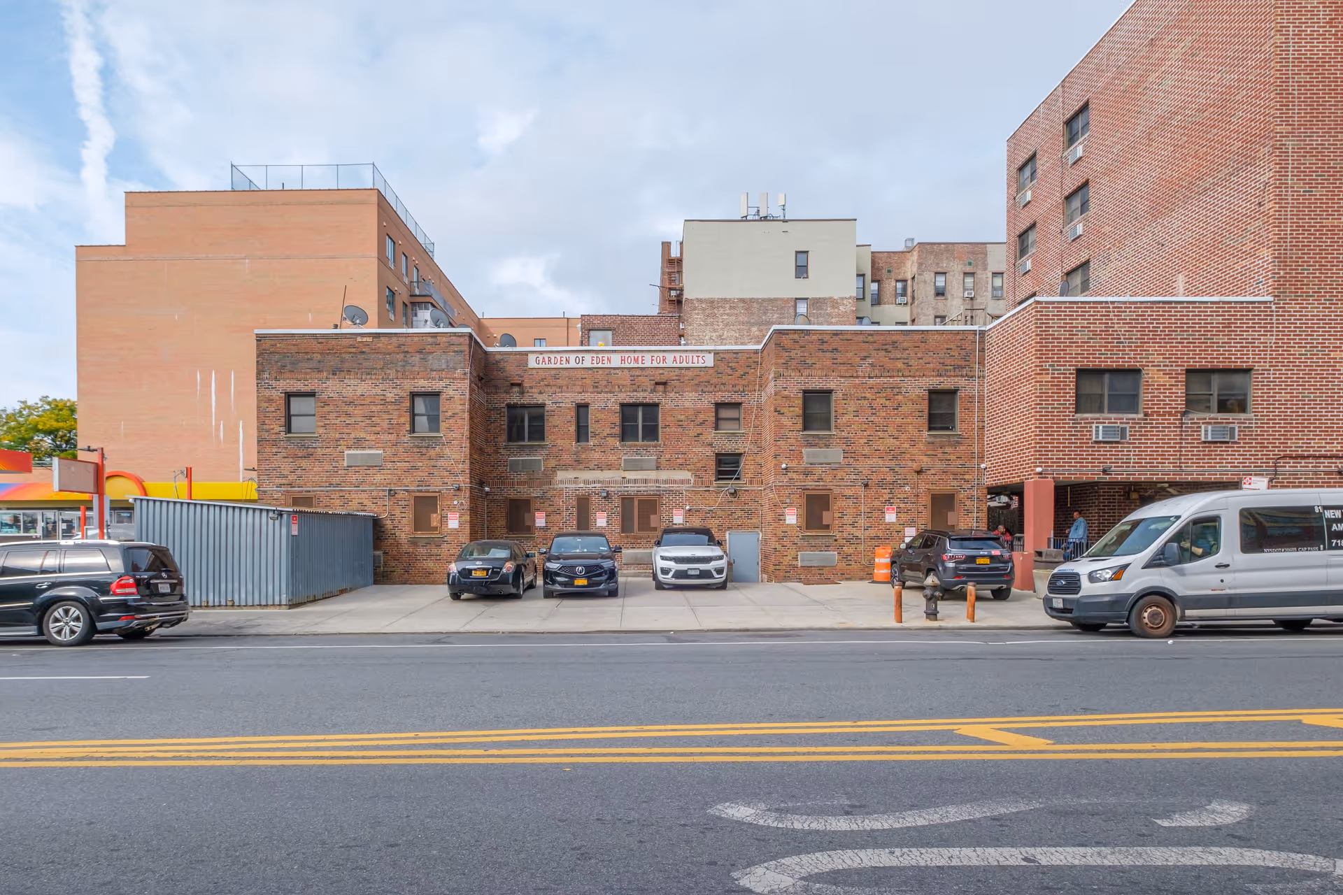 Front exterior view of the Garden of Eden Home for Adults building, a brick structure with several windows and a parking area in front with multiple parked cars. The building is surrounded by other taller buildings under a partly cloudy sky.