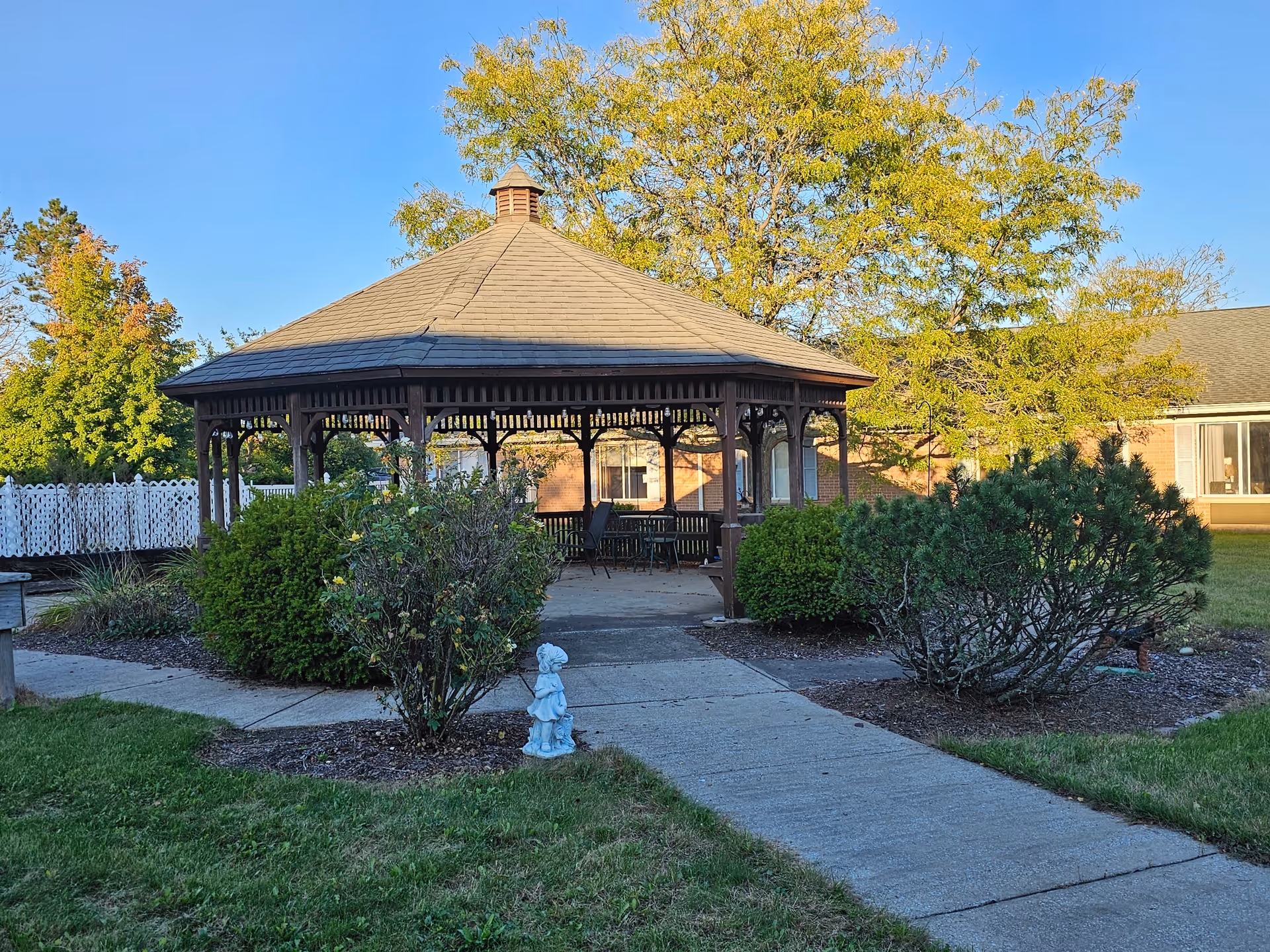 A wooden gazebo with a shingled roof situated in a garden area with bushes and a concrete pathway leading to it. There is a small statue of a child near the pathway. Trees and a building with large windows are visible in the background under a clear blue sky.
