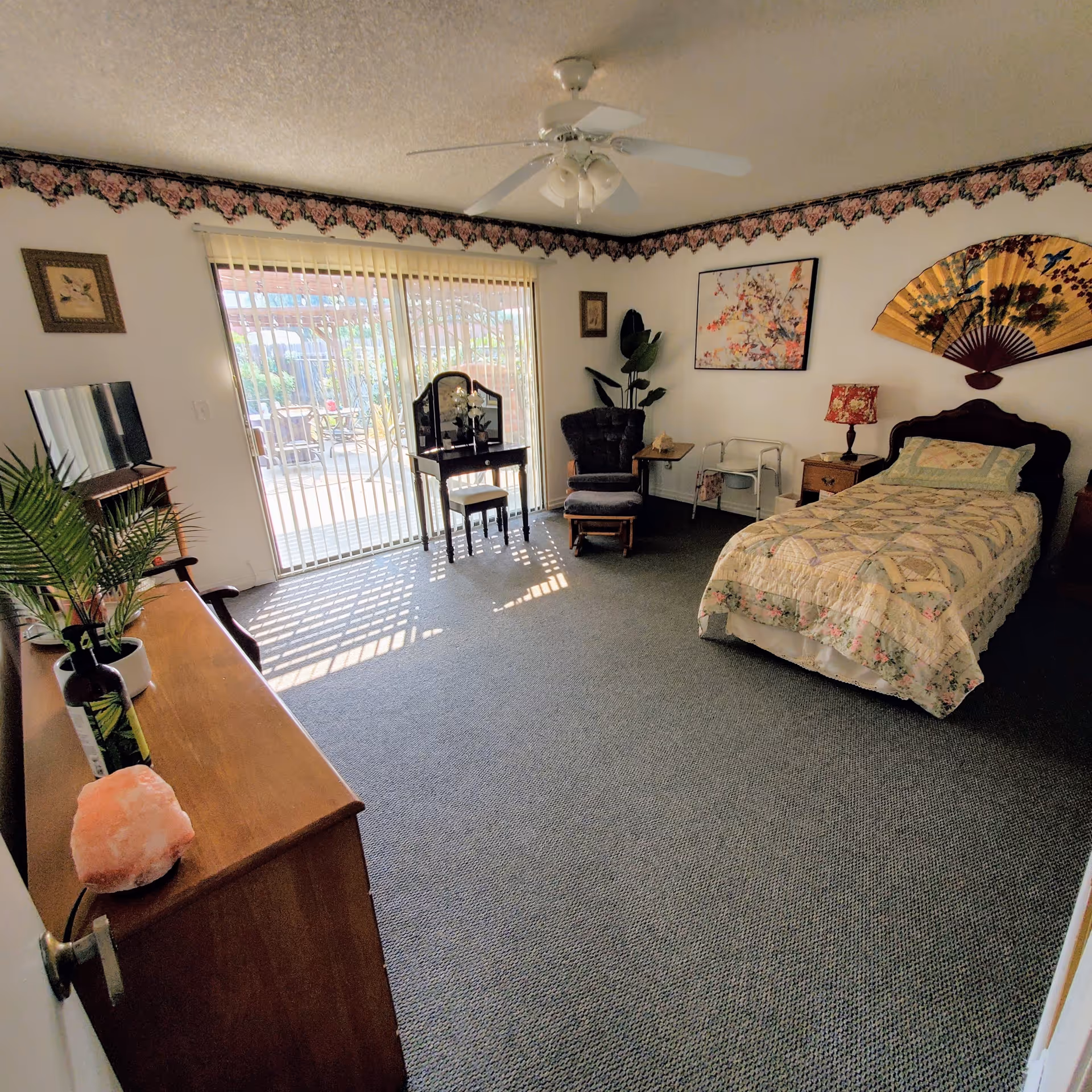 A cozy bedroom in a senior living facility with a single bed covered in a floral quilt, a wooden dresser with a TV on top, a small vanity table with a mirror and stool, a cushioned armchair with an ottoman, and sliding glass doors leading to an outdoor patio. The room has floral wallpaper border near the ceiling and decorative wall art including a large fan and framed pictures.