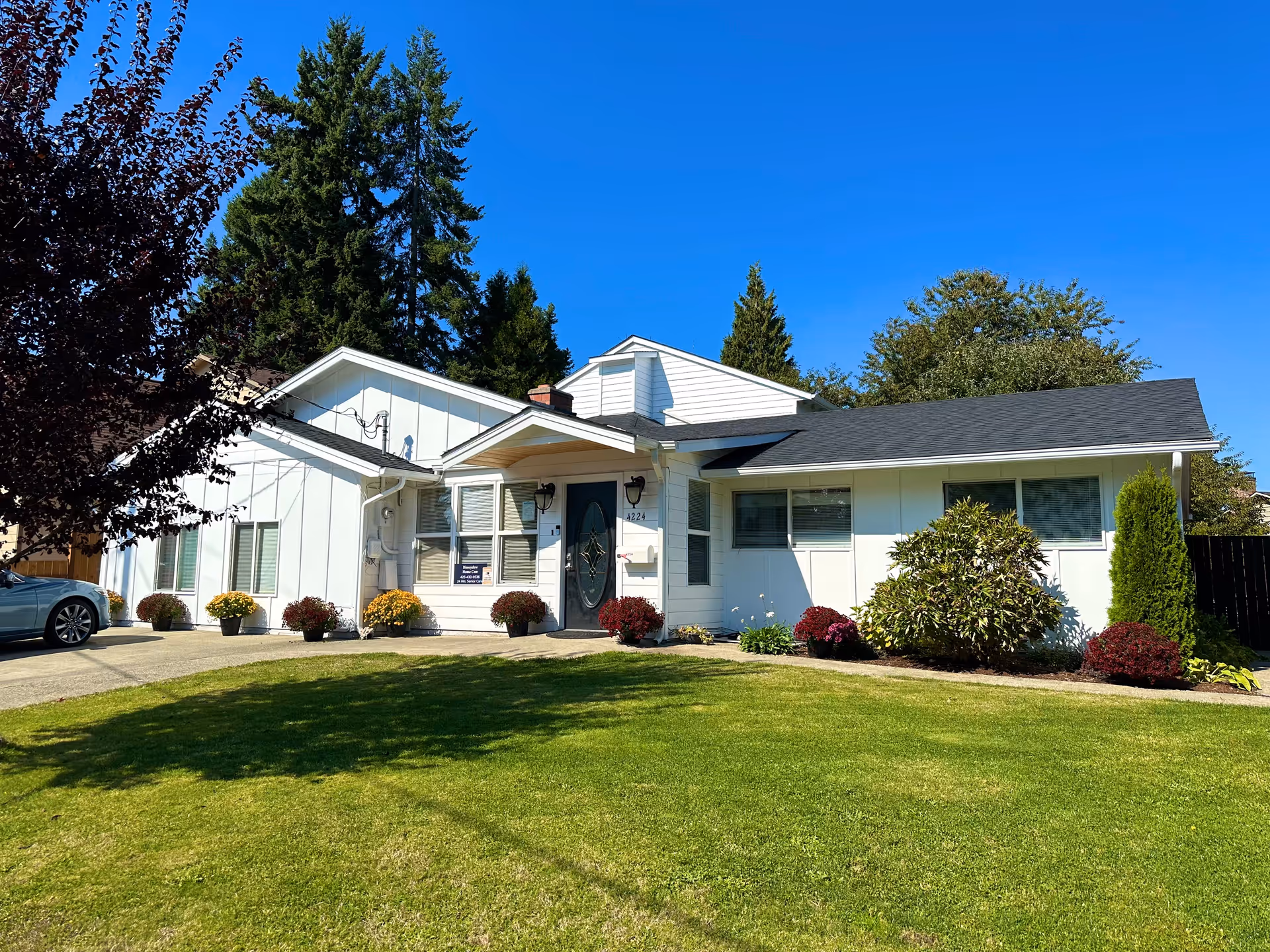 Front exterior of a single-story white house with a green lawn, potted flowers, and trees under a clear blue sky.