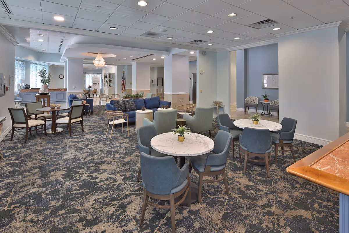 A spacious and well-lit common area in a senior living facility featuring multiple seating arrangements including round marble-top tables with blue upholstered chairs, a blue sofa with cushions, and additional chairs around small tables. The room has a patterned carpet, white walls, and ceiling lights. There is a reception desk area visible in the background along with an American flag and some decorative plants.