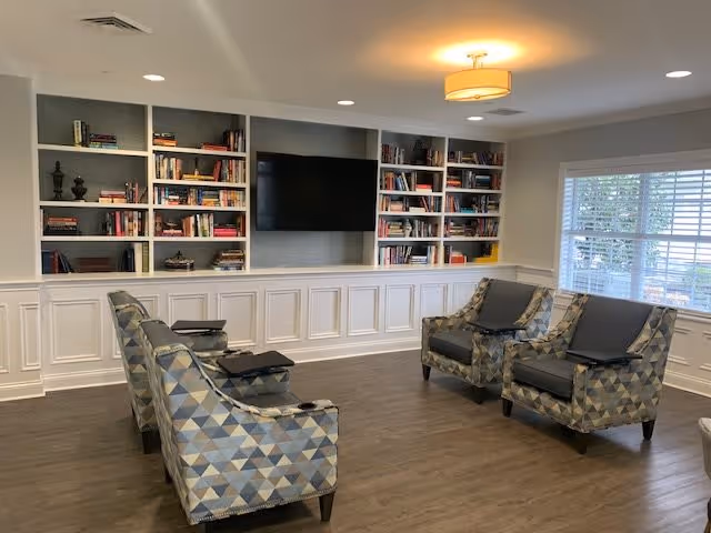 A cozy living room area in Chandler Park Assisted Living featuring patterned armchairs arranged in two rows facing a wall-mounted flat-screen TV. The wall behind the TV has built-in white bookshelves filled with books and decorative items. The room has wood flooring, white wainscoting, recessed lighting, a ceiling light fixture, and a window with white blinds allowing natural light in.