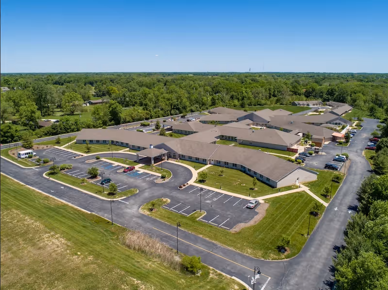 Aerial view of The Village Of Avon senior living facility showing a large, single-story building complex with multiple wings, surrounded by parking lots and green lawns, set against a backdrop of dense trees and clear blue sky.