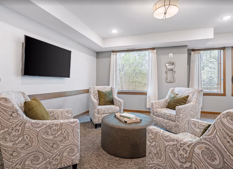 Cozy sitting room with four patterned armchairs arranged around a round ottoman, a wall-mounted TV, and windows with blinds.