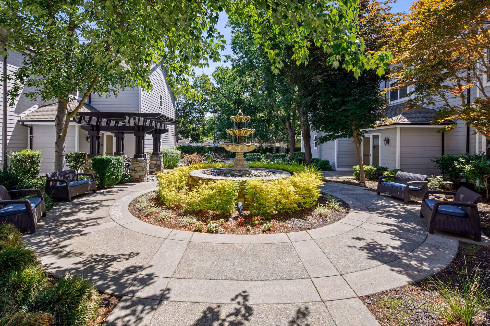 A peaceful outdoor courtyard area at Oakmont Of Brookside featuring a circular walkway surrounding a tiered stone fountain with yellow-green shrubs and various trees providing shade. There are several benches with cushions placed along the walkway, and light gray buildings with white trim border the courtyard.