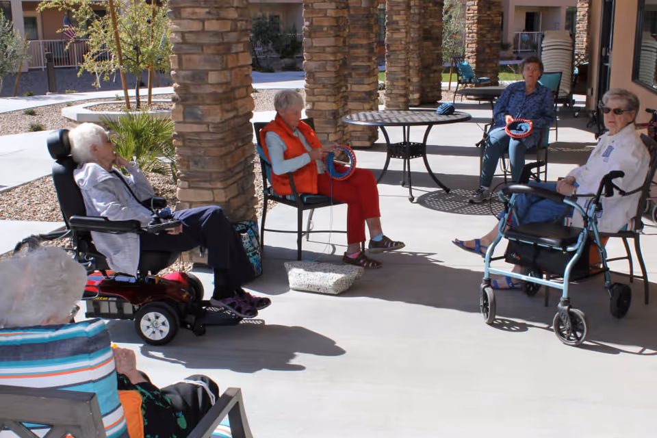 A group of elderly women sitting outside on a shaded patio area with stone pillars. Some women are seated in chairs, one is in a motorized wheelchair, and another has a walker nearby. Two women are holding circular knitting looms and appear to be knitting. There are tables and chairs around them, and the background shows part of the building and landscaping with small trees and gravel.