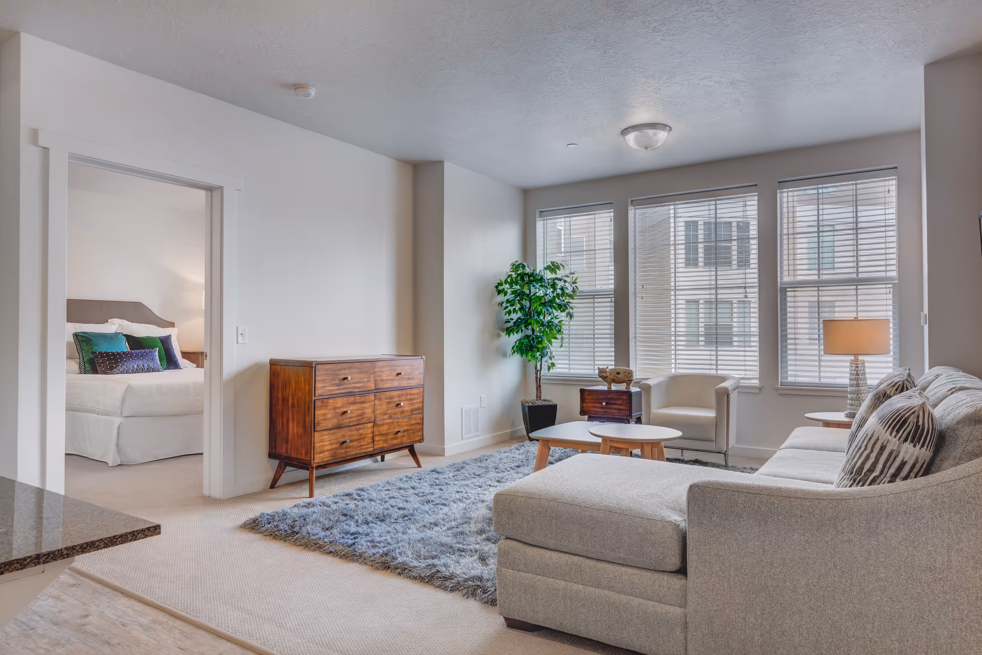 A bright and modern living room with a gray sectional sofa, a white armchair, two round wooden coffee tables, a wooden dresser, a green potted plant, and large windows with blinds. A bedroom with a bed and colorful pillows is visible through an open doorway.