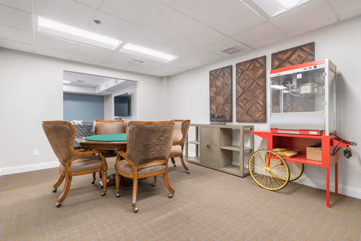 A room with a round table covered with a green felt top surrounded by four brown cushioned chairs with wooden legs on casters. To the right, there is a red and yellow popcorn machine on wheels. Behind the popcorn machine is a light wood cabinet with open shelves and three decorative wooden wall panels mounted above it. The room has beige carpet and white walls with a drop ceiling and fluorescent lighting.