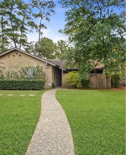 A single-story brick cottage with a pathway leading up to the entrance, surrounded by green grass, bushes, and tall trees under a blue sky.