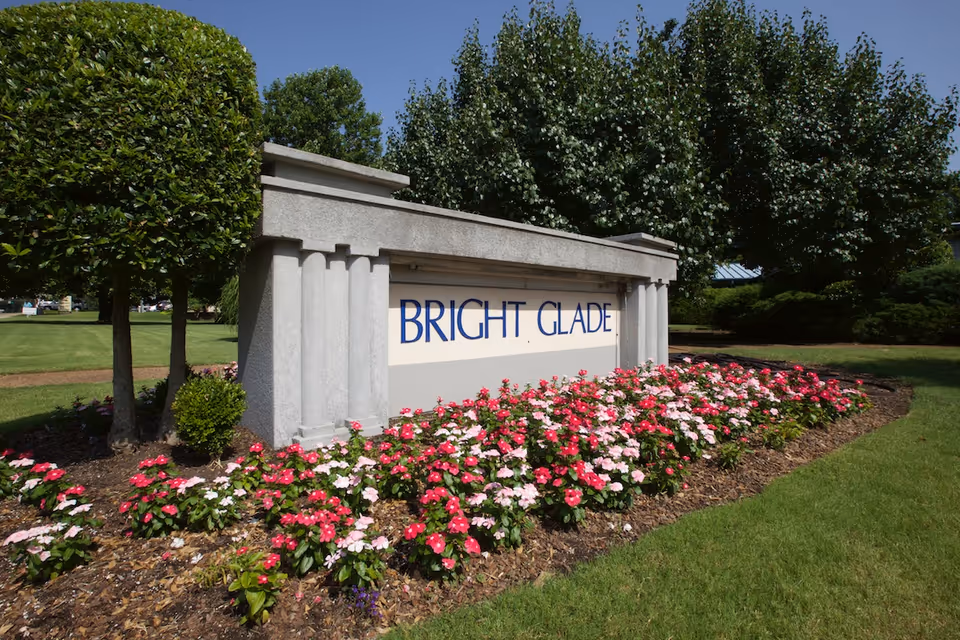 Stone sign with the words 'BRIGHT GLADE' surrounded by a flower bed with pink and red flowers, green grass, and trees in the background under a clear blue sky.