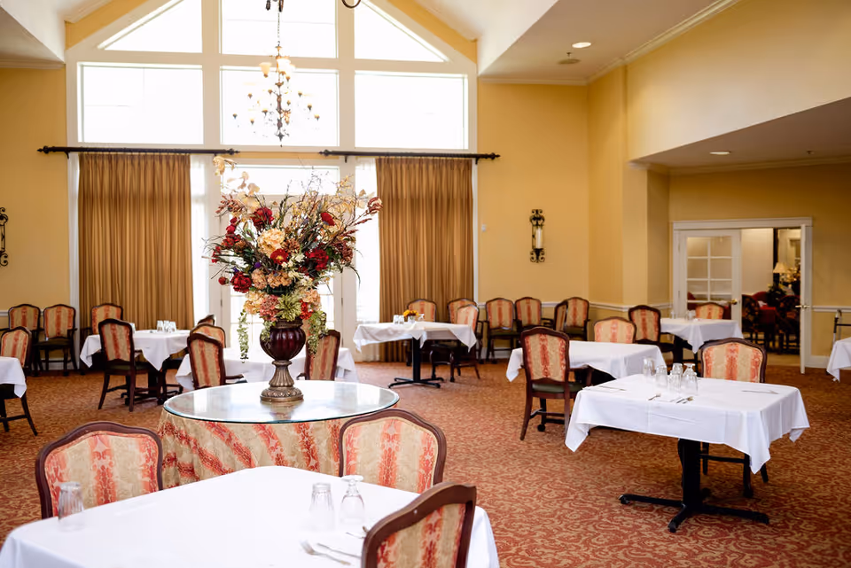 A spacious dining room with multiple tables covered in white tablecloths and set with glasses and napkins. The chairs have a classic design with red and beige upholstery. A large floral arrangement in a vase is placed on a round table in the center. The room features high ceilings with large windows and beige curtains, and a chandelier hanging from the ceiling.