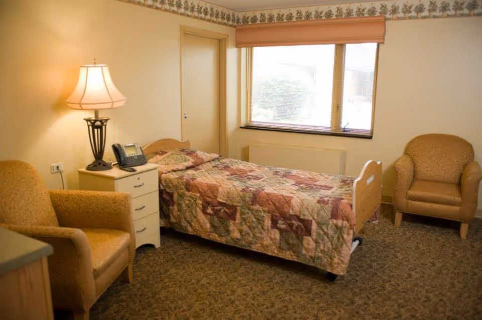 A small, cozy bedroom with a single bed covered in a patchwork quilt. There are two upholstered armchairs, one on each side of the room. A nightstand with a lamp and a telephone is next to the bed. A large window with blinds lets in natural light.