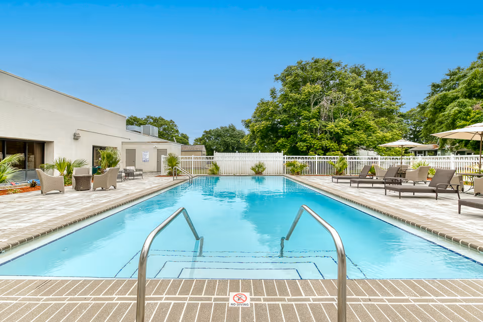 Outdoor swimming pool with clear blue water surrounded by a tiled deck. There are lounge chairs and umbrellas on the right side and wicker chairs with a small table on the left side near a building. Green trees and a white fence are visible in the background under a clear blue sky.