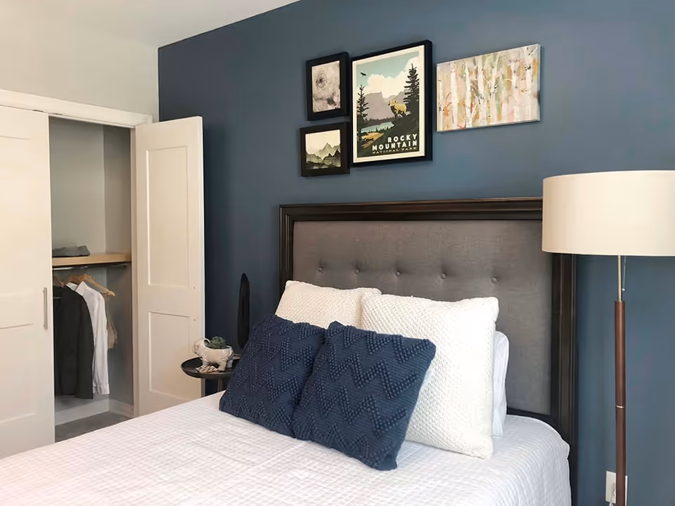 Bedroom with a bed dressed in white linens and blue pillows, a tufted headboard, wall art above, an open closet to the left, and a floor lamp to the right.