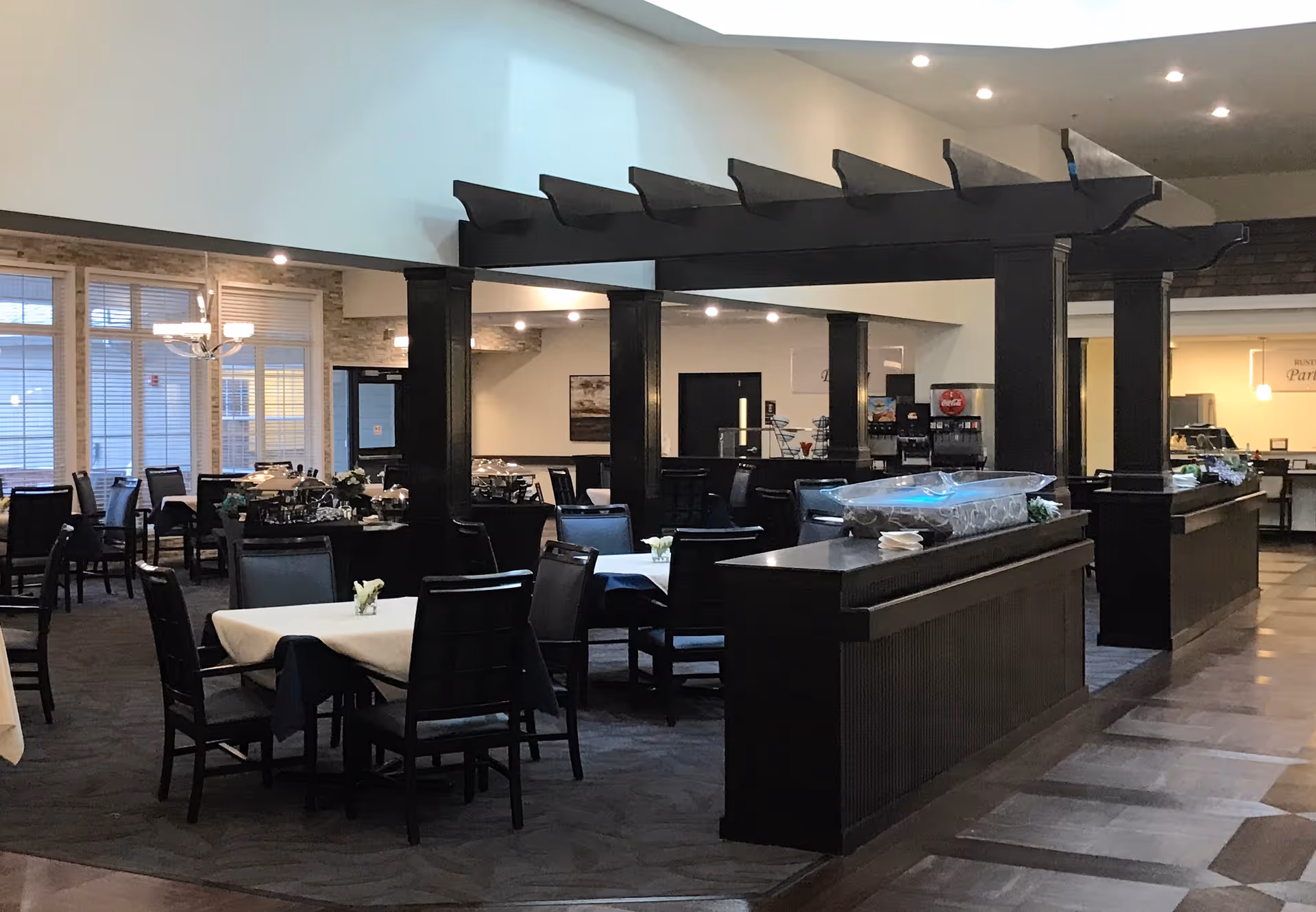 Interior view of a dining room in a senior living facility with multiple tables and chairs arranged neatly. The room features dark wooden furniture, a decorative overhead beam structure, and large windows with blinds allowing natural light. There is a buffet or serving area with covered trays and a beverage station in the background.
