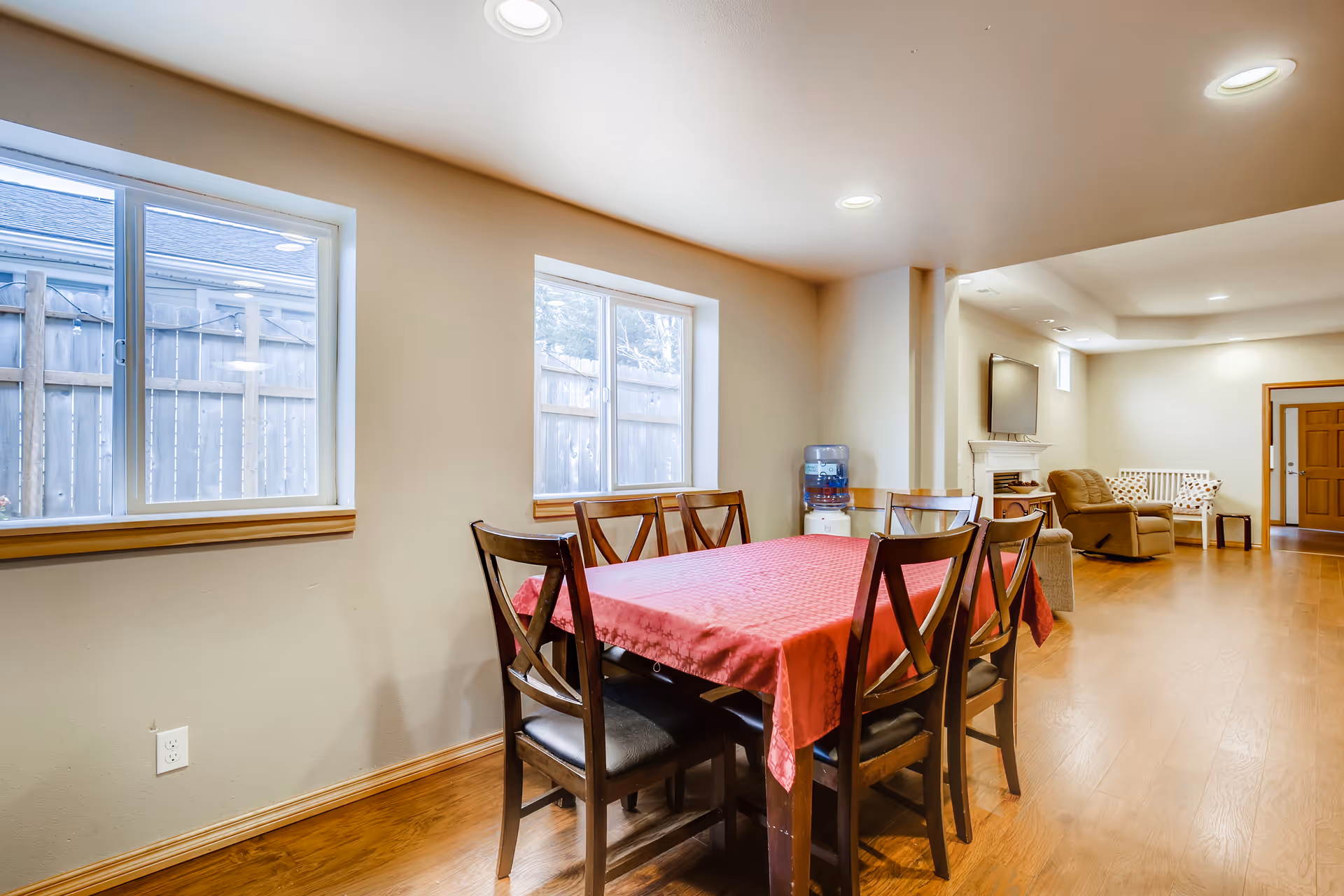 Interior view of a senior living facility dining and living area with a wooden dining table covered with a red tablecloth and six chairs. Two windows with a view of a wooden fence let in natural light. In the background, there is a water dispenser, a beige recliner chair, a white bench with patterned cushions, a small side table, and a wall-mounted flat-screen TV above a white fireplace. The room has wooden flooring and recessed ceiling lights.