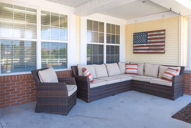 Covered outdoor patio area with a wicker sectional sofa and a matching armchair, both with beige cushions and red-and-white striped throw pillows. An American flag decoration is mounted on the yellow siding wall behind the sofa. The area has large windows with white blinds and a brick half-wall.