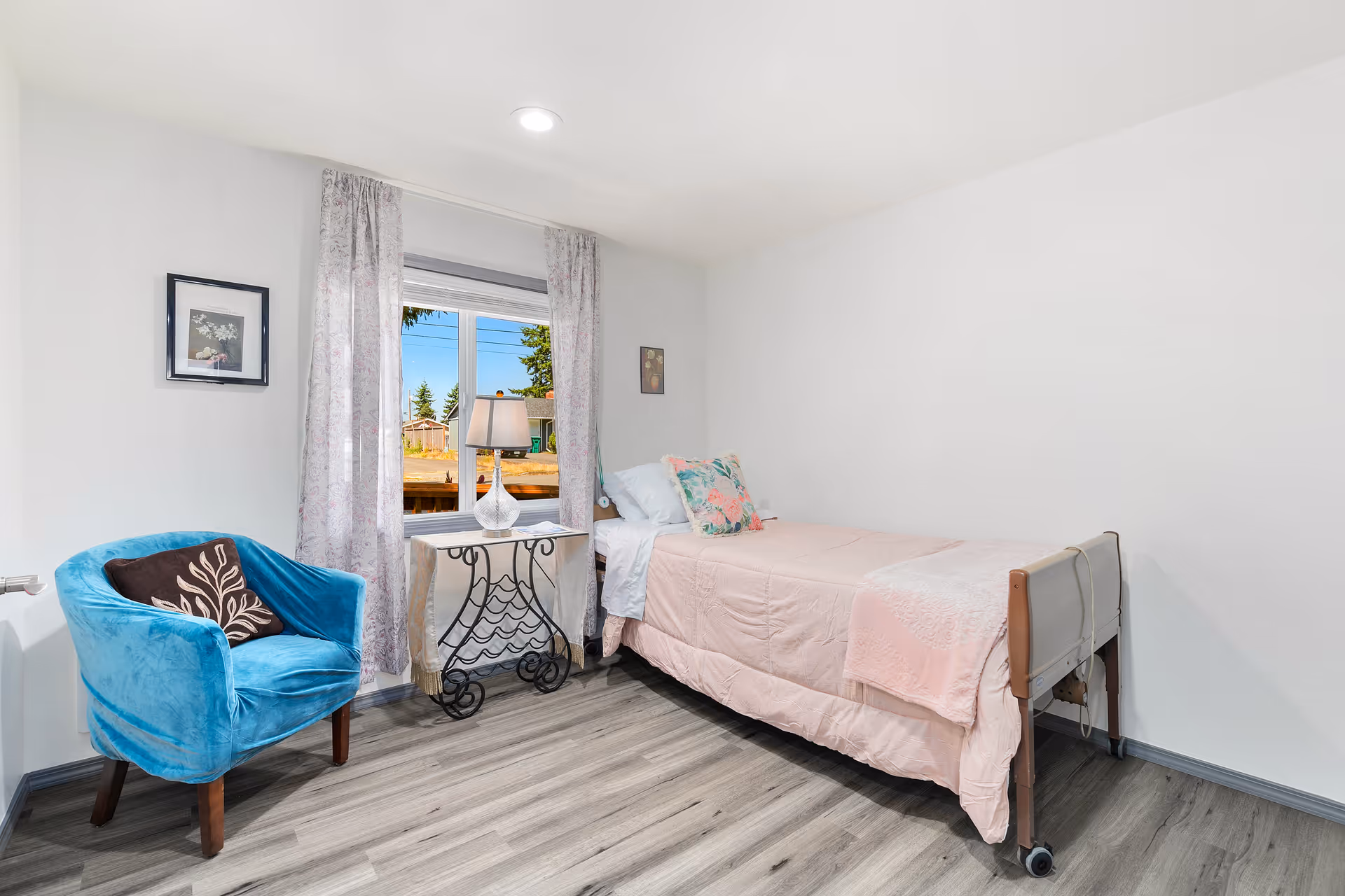 A small, bright bedroom with a single bed covered in light pink bedding and floral pillows. Next to the bed is a decorative metal table with a lamp on it, positioned in front of a window with sheer curtains. A blue upholstered chair with a brown pillow is placed near the window. The room has light-colored walls and wood-patterned flooring.