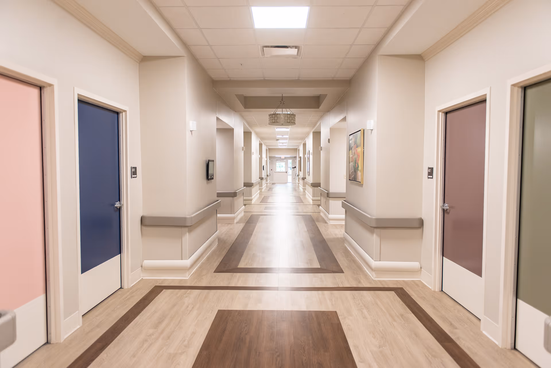 Well-lit long corridor in a senior living facility with colorful doors and handrails along the walls.