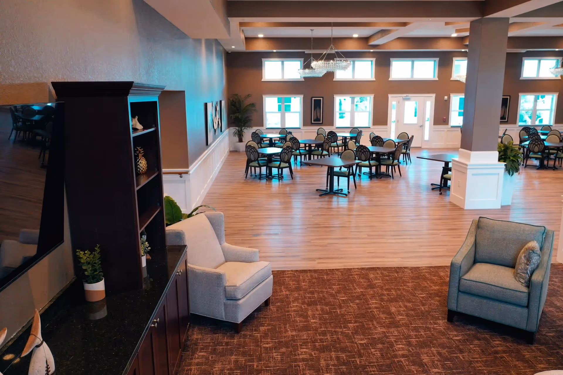 Spacious dining area with multiple round tables and chairs arranged on a wooden floor, large windows letting in natural light, and two comfortable armchairs in the foreground on a carpeted area next to a dark wooden cabinet with decorative items.