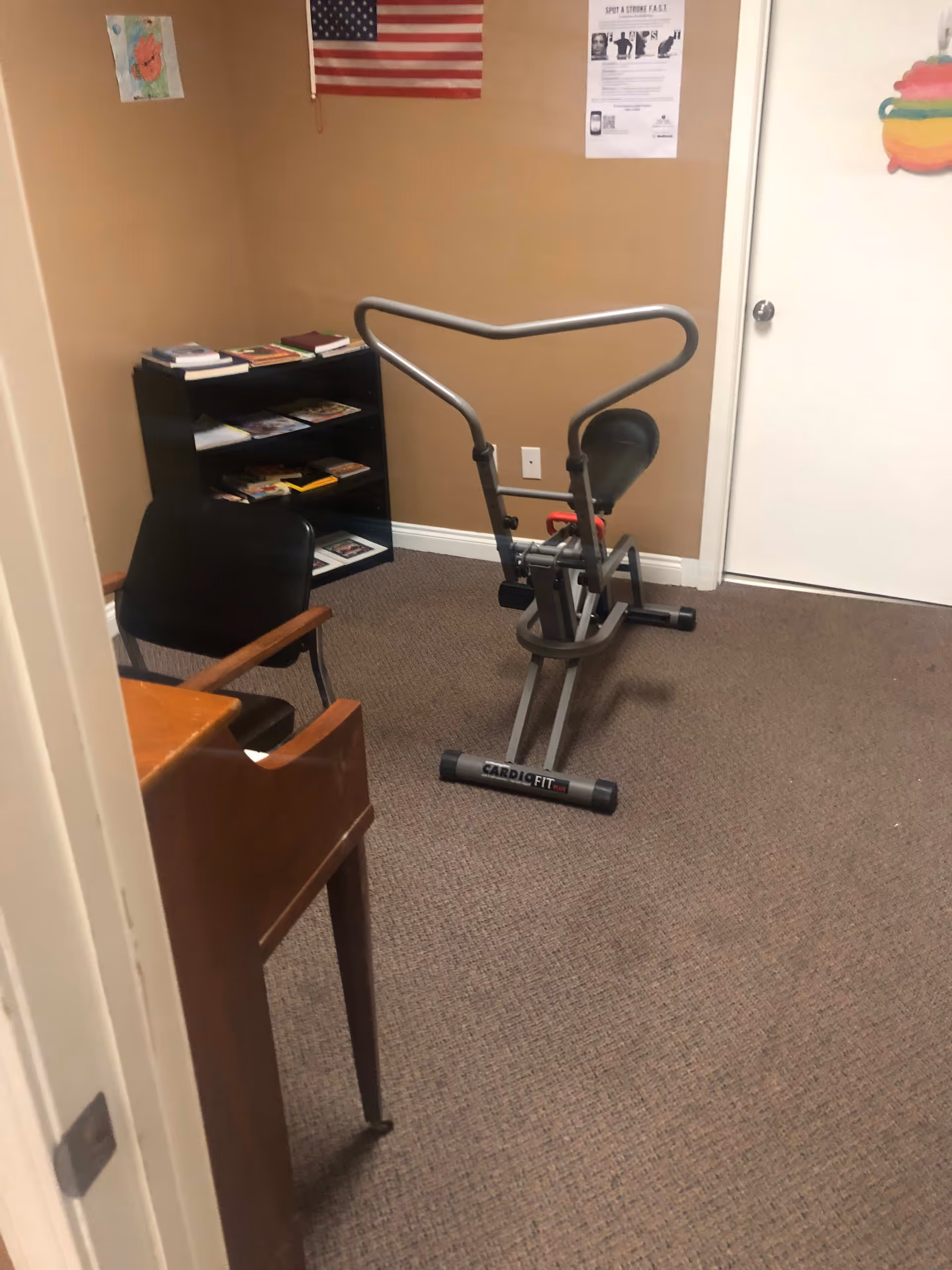 A small room with beige walls and carpeted floor containing a stationary exercise bike labeled 'Cardio Fit', a black chair with wooden armrests, a wooden desk, and a black bookshelf filled with books and magazines. An American flag and some posters are hung on the walls.