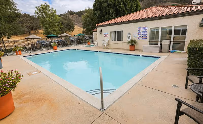 Outdoor rectangular swimming pool with a concrete deck, patio chairs and umbrellas beside a single-story building with a tiled roof and hills in the background.