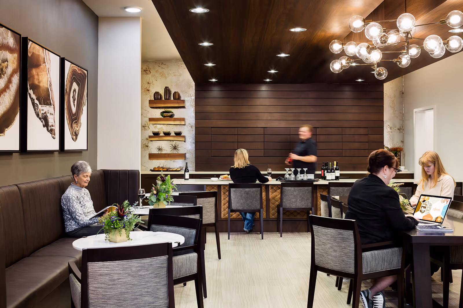 Residents and staff sit at tables and a bar in a modern communal lounge with a wood-paneled ceiling and decorative lighting.
