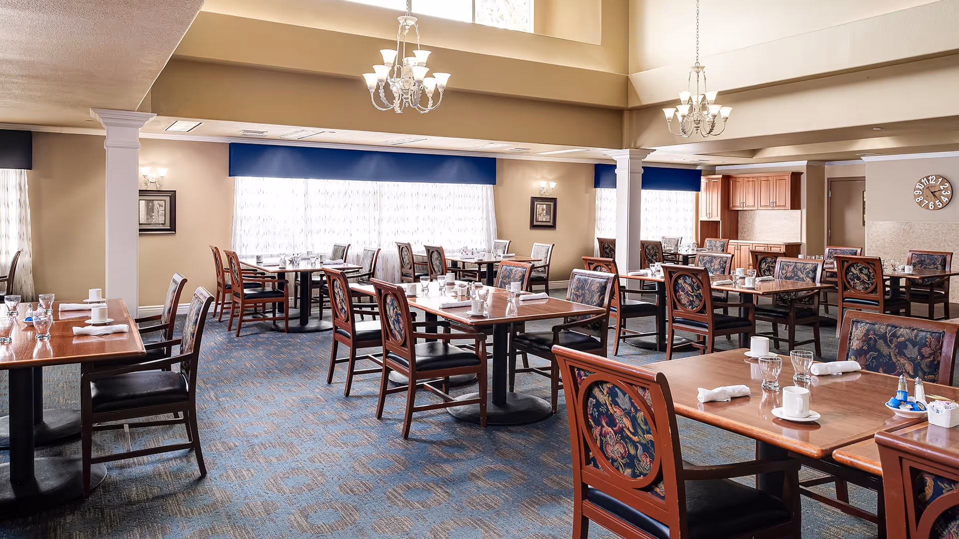 A spacious dining room with multiple wooden tables and chairs arranged neatly. Each table is set with white cups, glasses, and napkins. The room features large windows with sheer curtains and blue valances, beige walls, carpeted floor with a patterned design, and two chandeliers hanging from the ceiling.