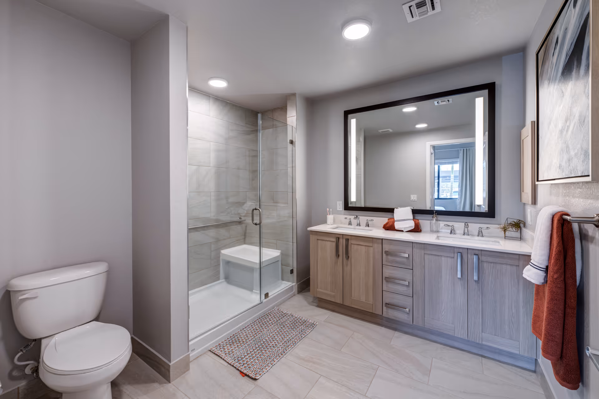 Modern bathroom with a white toilet, a glass-enclosed shower with a built-in bench, a large mirror with built-in lights above a double sink vanity with wooden cabinets, and towels hanging on a rack.