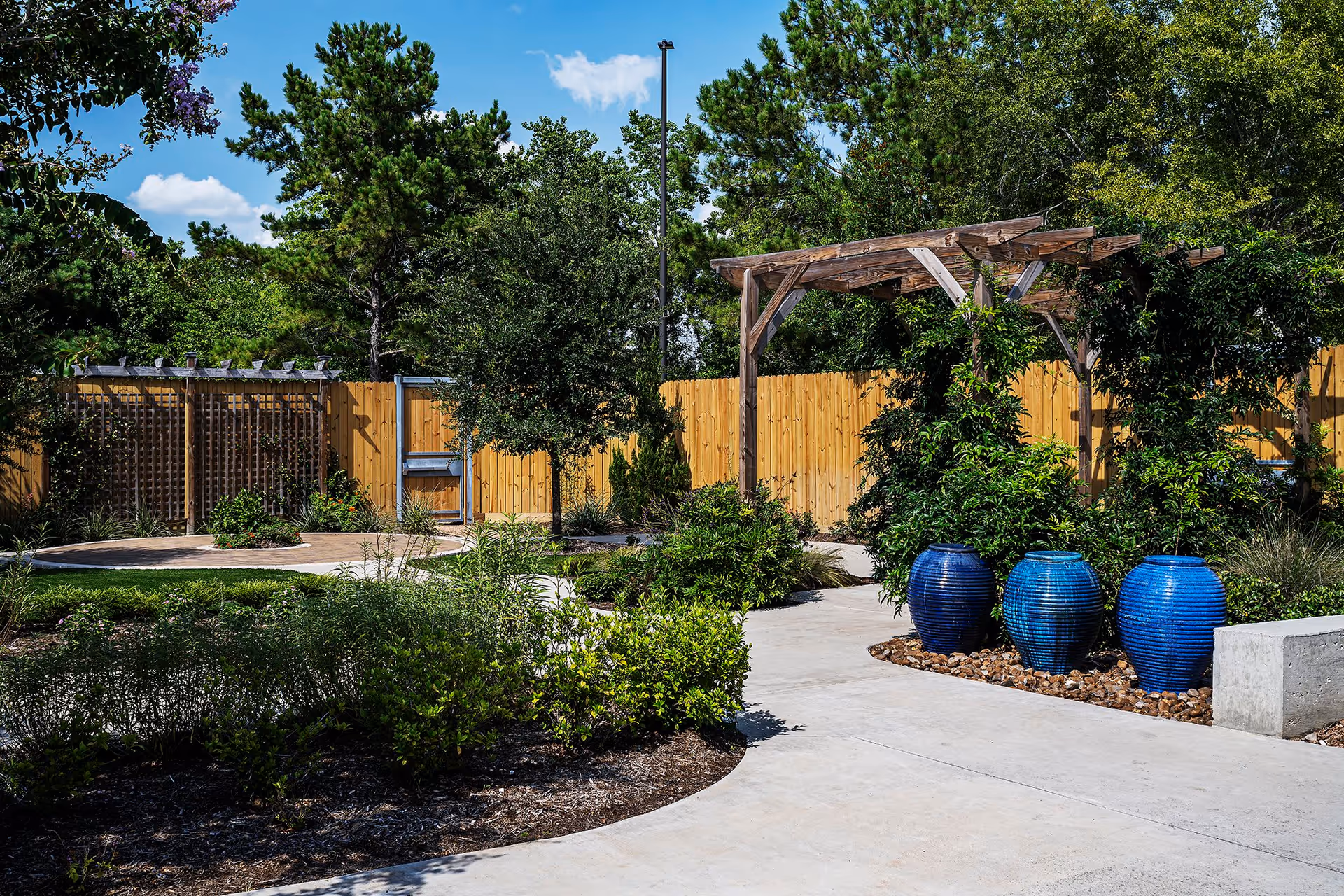 Outdoor courtyard garden with a wooden pergola, three large blue ceramic planters, pathways, and landscaped greenery.