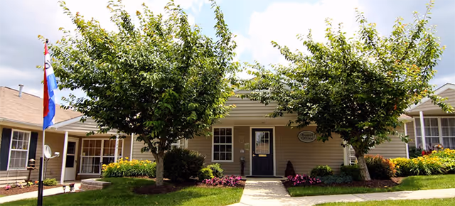 Exterior view of a single-story building with beige siding, a dark front door, and two large leafy trees in front. There is a paved walkway leading to the door, flower beds with colorful flowers on either side, and a flagpole with a flag on the left side of the image. The sky is partly cloudy.