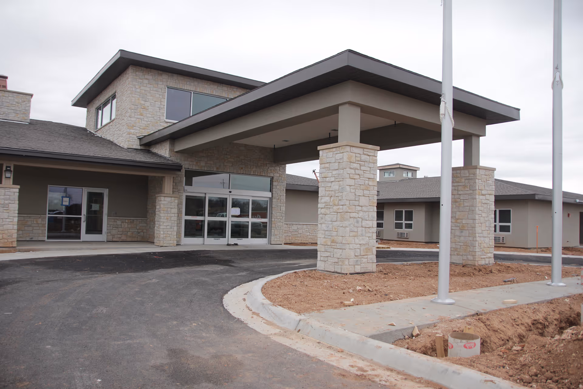 Exterior view of a modern senior living facility entrance with stone pillars supporting a covered drop-off area. The building has large windows and a paved driveway with some landscaping work in progress.