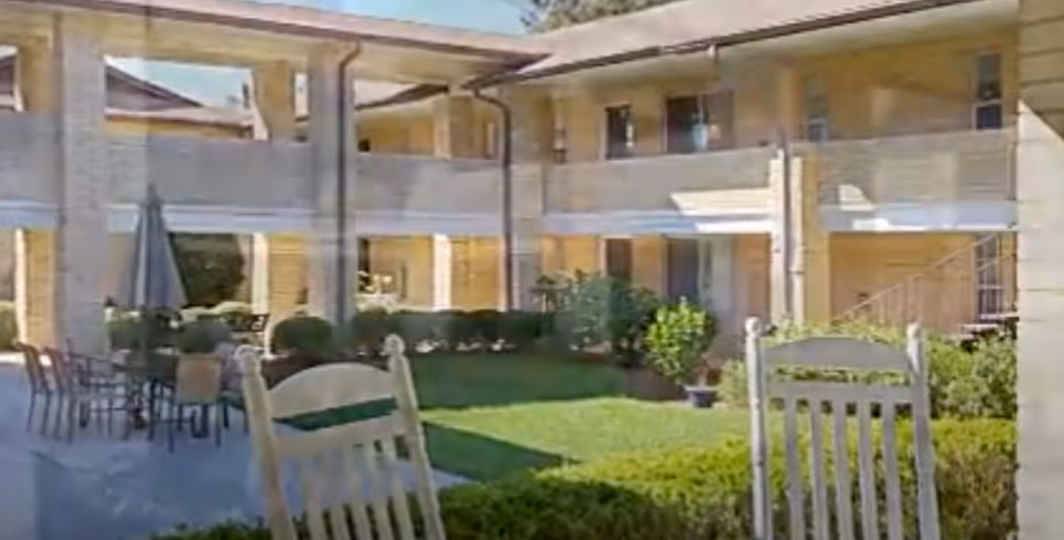 Outdoor courtyard area of Camelot Chateau Assisted Living featuring green grass, bushes, patio tables with chairs, and a two-story building with balconies surrounding the courtyard.