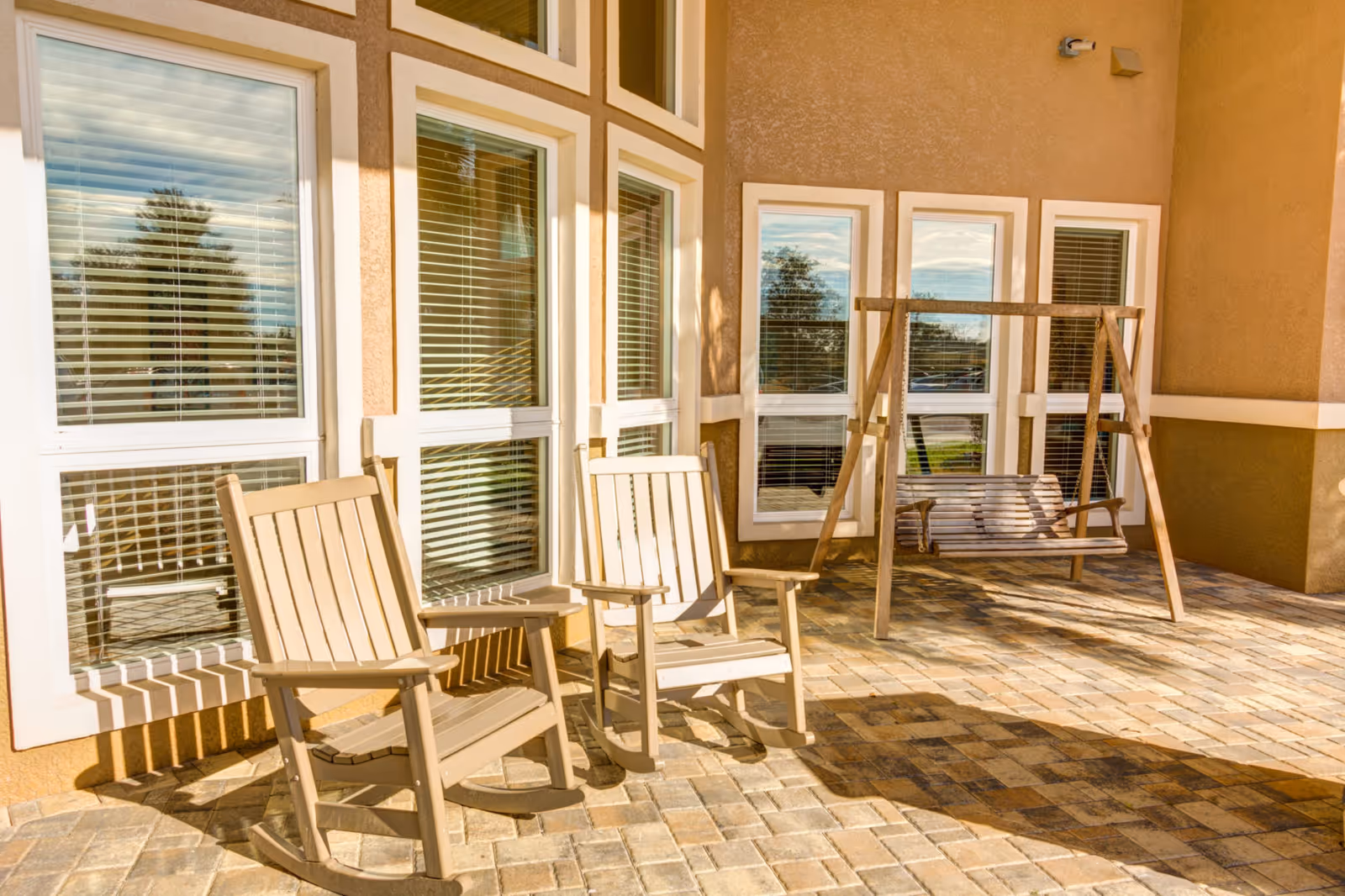 Outdoor patio area with two wooden rocking chairs and a wooden swing bench in front of a building with multiple windows and beige walls.