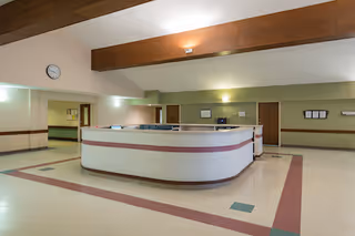 Interior view of a spacious reception area in a senior living facility with a large curved white reception desk with a pink stripe, beige and green tiled floor, light green walls, wooden doors, and a clock on the wall.