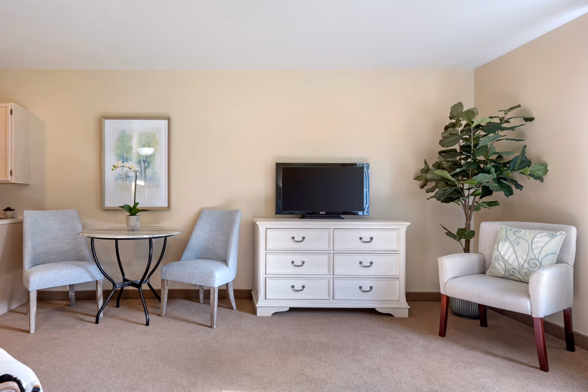 A cozy living area with a small round table and two light gray upholstered chairs on the left, a white dresser with six drawers and a flat-screen TV on top in the center, and a light gray armchair with a decorative pillow next to a tall green potted plant on the right. The walls are beige and there is a framed artwork hanging above the table.