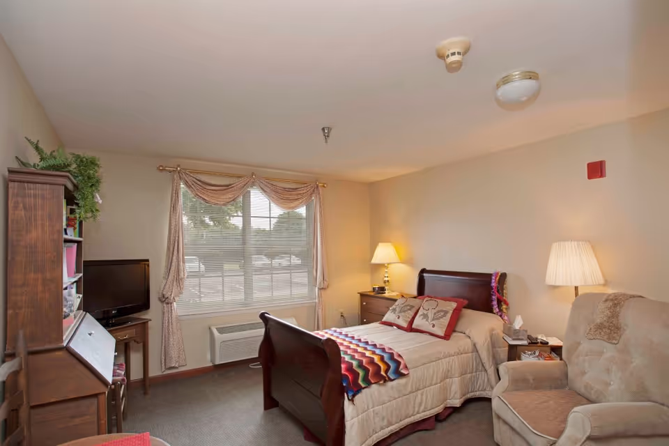 A cozy bedroom in a senior living facility with a wooden bed featuring a beige comforter and decorative pillows. There is a colorful blanket draped at the foot of the bed. Next to the bed is a nightstand with a lamp and tissues. A comfortable armchair with a throw blanket is positioned nearby. A window with beige curtains allows natural light into the room. A wooden desk with shelves and a small TV on a stand are also visible.