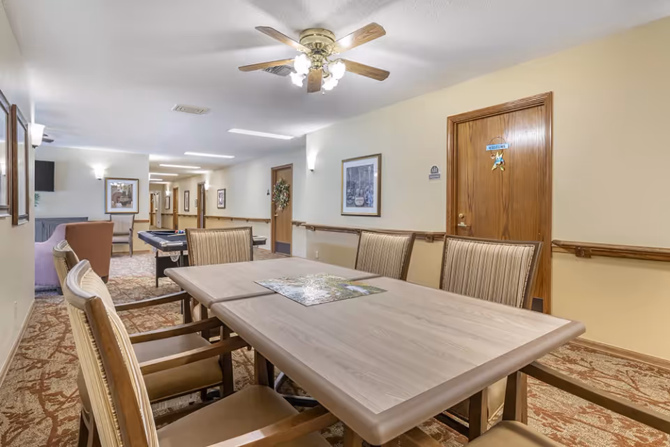 A well-lit interior room with a rectangular table surrounded by six chairs. On the table is a partially completed jigsaw puzzle. The room has beige walls, carpeted floors with a patterned design, and a ceiling fan with lights. In the background, there is a pool table and some seating areas along a hallway with several doors and framed pictures on the walls.