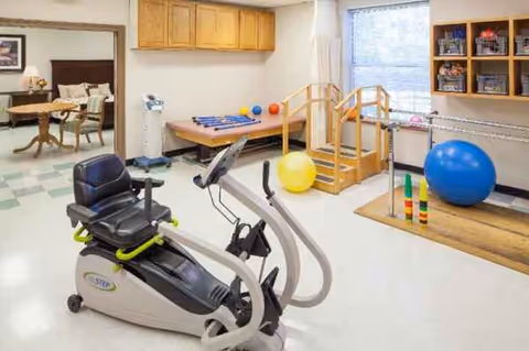 Interior rehab room at a senior facility with exercise bike, stairs, balls, and shelves.