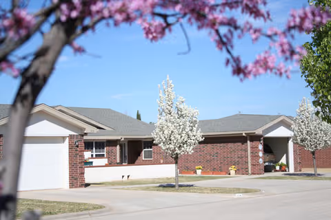 Single-story brick senior living units with garages, driveways, and blossoming trees in the foreground.