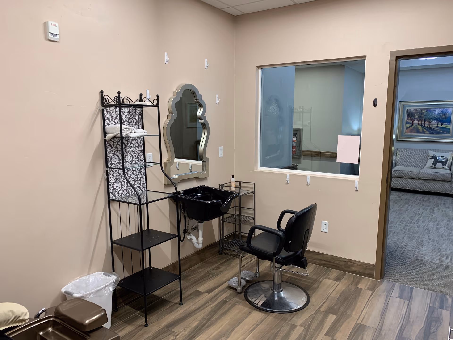 Interior view of a small salon or grooming area with a black salon chair in front of a black sink and a decorative mirror mounted on a beige wall. Next to the sink is a black metal shelving unit with towels and other items. The floor has wood-like tiles and there is a window looking into another room with a couch and a framed picture on the wall.