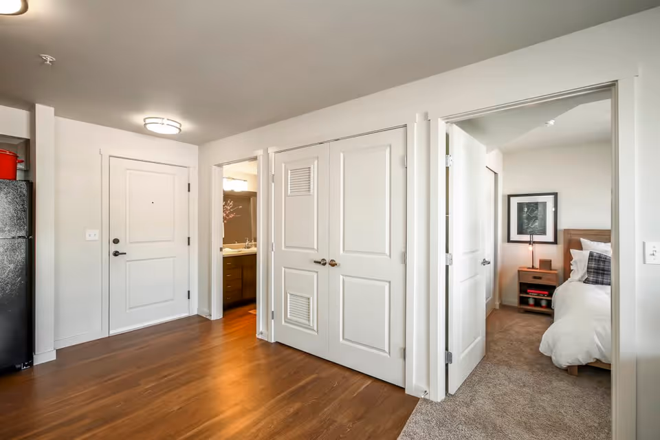 Interior view of a senior living facility apartment at Vintage at Mill Creek showing a hallway with wooden flooring, a white entrance door, a closet with double doors, an open door leading to a bathroom with a sink and mirror, and another open door leading to a bedroom with a bed, nightstand, lamp, and framed artwork on the wall.
